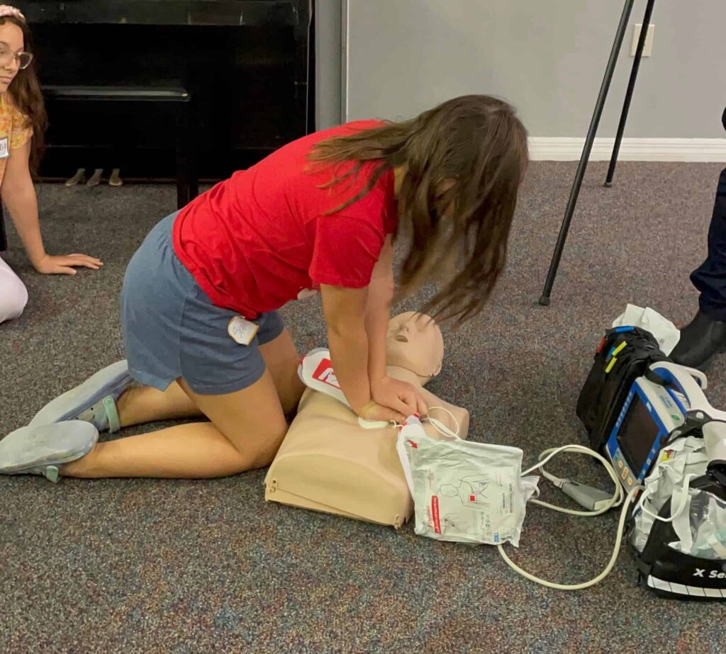 Young female student in red t-shirt and shorts kneeled down and practicing cpr on dummy.